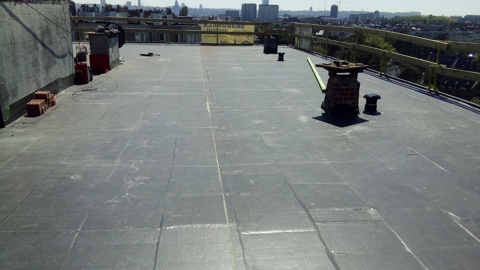 Płaski dach pokryty papą termozgrzewalną z widocznymi łączeniami, komin wentylacyjny i ceglany komin z deską opartą o niego. W tle panorama miejska.