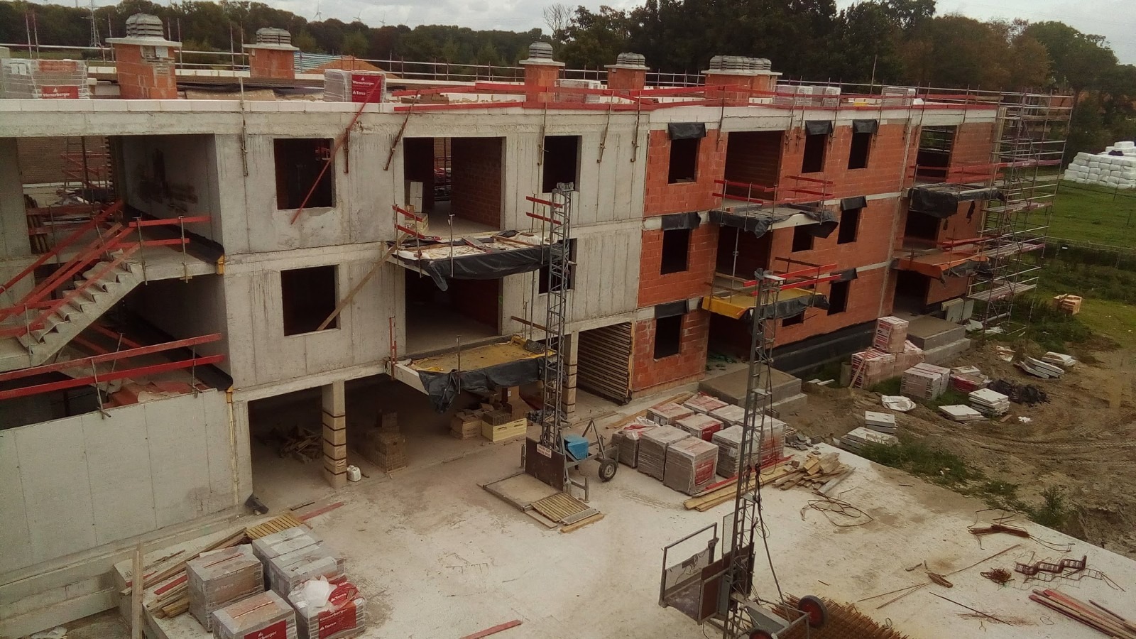 Wznoszenie trzypiętrowego budynku z betonu i czerwonej cegły, widoczne rusztowania, windy budowlane i materiały budowlane, w tle drzewa.