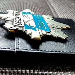 Metalowa odznaka z napisem 'Detektyw' i niebieskim akcentem, leżąca na czarnym skórzanym portfelu na tle szarej tkaniny.
