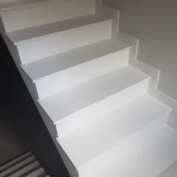 Nowoczesne, pomalowane na biało schody wewnętrzne z widocznym fragmentem schodów prowadzących w dół, z delikatnym oświetleniem padającym z góry, ukazujące minimalistyczny design.