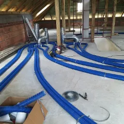 Instalacja systemu wentylacji z niebieskich, karbowanych rur w surowym pomieszczeniu na poddaszu, widoczne elementy metalowe i narzędzia montażowe.