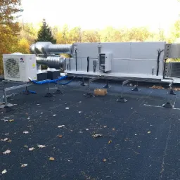 System wentylacyjny z klimatyzatorem na dachu budynku, widoczne kanały wentylacyjne, jednostka zewnętrzna klimatyzacji marki Gree, tło z drzewami w jesiennych barwach.