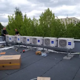 Rząd zewnętrznych jednostek klimatyzacyjnych Haier ustawionych na dachu budynku, widoczni dwaj instalatorzy podczas pracy, w tle drzewa i pochmurne niebo.