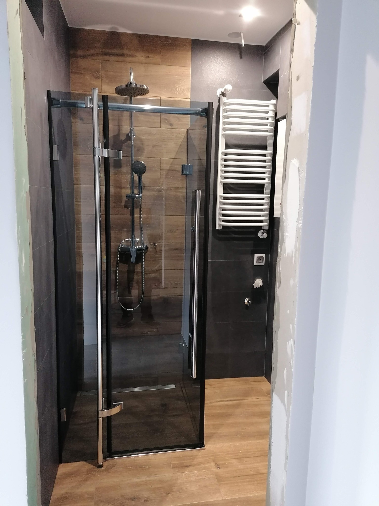 Nowoczesna łazienka w trakcie wykończenia: kabina prysznicowa z czarnymi ramami, grzejnik drabinkowy na ciemnoszarych płytkach, drewnopodobne płytki na ścianie prysznica, widoczne ślady prac...
