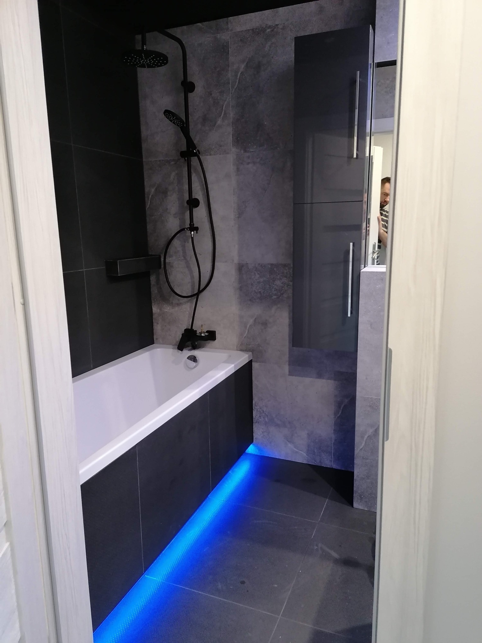Nowoczesna łazienka z czarno-szarymi płytkami imitującymi marmur, białą wanną z czarną obudową i czarną armaturą, podświetleniem LED w kolorze niebieskim pod wanną oraz wysoką szafką z lustrem.