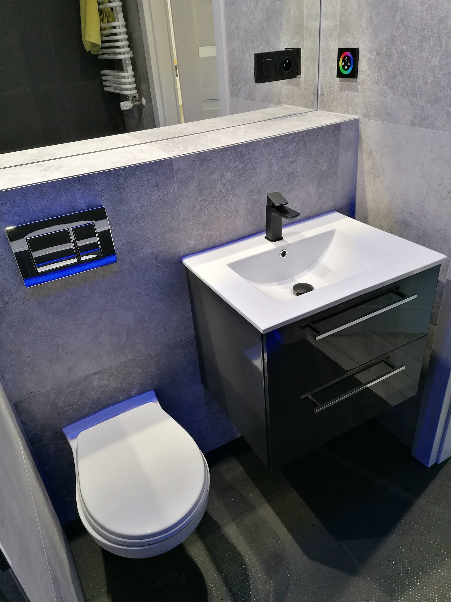 Nowoczesna łazienka z szarymi płytkami, podwieszaną szafką z umywalką, czarną armaturą i podświetlanym panelem sterowania.