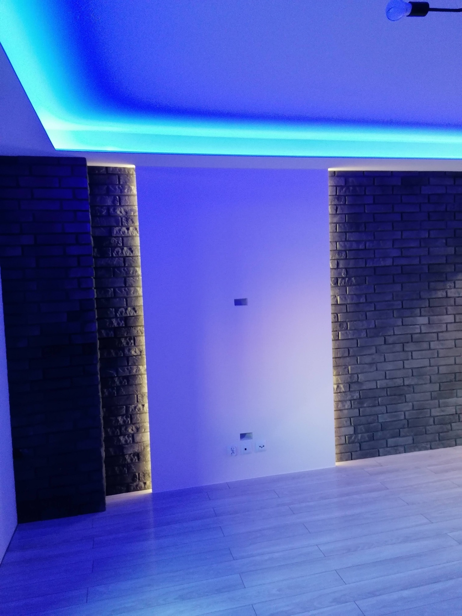 Nowoczesne wnętrze z ceglaną ścianą, podświetlanym sufitem i panelami podłogowymi, oświetlone niebieskim światłem.