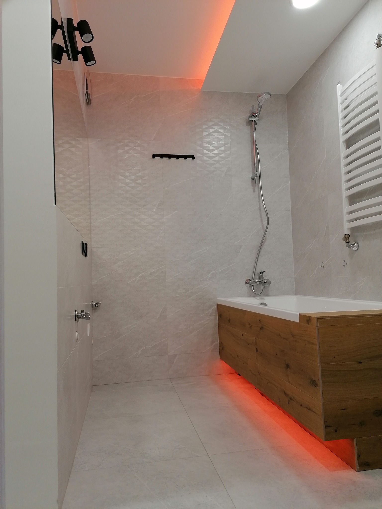 Nowoczesna łazienka z wanną obudowaną drewnopodobną okładziną, podświetloną od dołu czerwonym światłem LED, oraz jasnymi, marmurkowymi płytkami na ścianach i podłodze, uzupełniona o grzejnik...