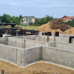 Wylany betonowy fundament domu jednorodzinnego z wystającymi prętami zbrojeniowymi, widoczne czarne szalunki, w tle koparka i zabudowa jednorodzinna.
