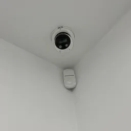 Kamera monitoringu BCS i czujnik ruchu zamontowane w rogu pomieszczenia z białymi ścianami i sufitem.