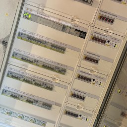 JLN Technology Systems Dariusz Zbigniew Urbański - Rozbudowana szafa elektryczna z modułami sterowania oświetleniem i roletami, widoczne liczne bezpieczniki, bloki rozdzielcze i sterowniki z wyświetlaczami LED w białej obudowie, instalacja...