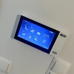 Panel dotykowy wideodomofonu BCS zamontowany na białej ścianie, widoczny ekran z ikonami i datą 24-10-2022, obok włączniki światła.