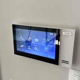 JLN Technology Systems Dariusz Zbigniew Urbański - Nowoczesny wideofon BCS z dotykowym ekranem, zamontowany na białej ścianie obok włącznika światła.