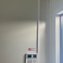 JLN Technology Systems Dariusz Zbigniew Urbański - Biała ściana z zamontowanym panelem sterowania alarmem, czujnikiem ruchu pod sufitem i naklejką firmy instalującej systemy zabezpieczeń.