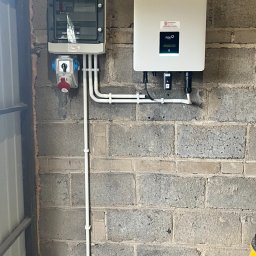 JLN Technology Systems Dariusz Zbigniew Urbański - Rozdzielnia elektryczna i inwerter fotowoltaiczny zamontowane na szarej, betonowej ścianie z widocznymi przewodami w białych rurkach PCV.