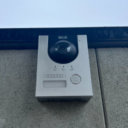 JLN Technology Systems Dariusz Zbigniew Urbański - Srebrny wideodomofon BCS zamontowany na szarej, betonowej ścianie zewnętrznej budynku, widok z dołu.