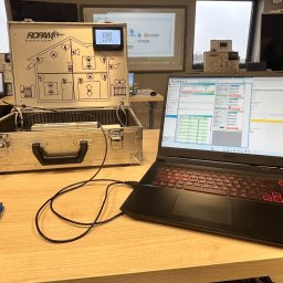 JLN Technology Systems Dariusz Zbigniew Urbański - Laptop z uruchomionym oprogramowaniem do konfiguracji systemu alarmowego, obok walizka z panelem sterującym i schematem instalacji, widoczni ludzie w tle.