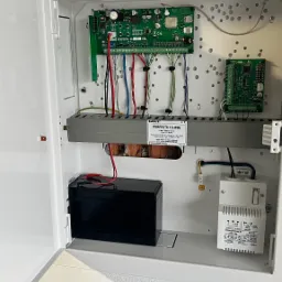 Otwarta skrzynka z elektroniką alarmową marki Satel Perfecta 16-WRL, akumulatorem i transformatorem, widoczne kable i płytki drukowane.