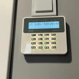 Biały panel sterowania systemu alarmowego Satel Perfecta z podświetlanymi przyciskami, zamontowany na szarej ścianie obok włącznika światła.