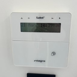 JLN Technology Systems Dariusz Zbigniew Urbański - Biały panel sterowania systemu alarmowego Satel Integra z wyświetlaczem LCD pokazującym datę i godzinę, zamontowany na białej ścianie obok czarnego włącznika światła.