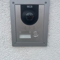 JLN Technology Systems Dariusz Zbigniew Urbański - Szary wideofon BCS zamontowany na białej, chropowatej ścianie zewnętrznej budynku, widoczne przyciski i głośnik.