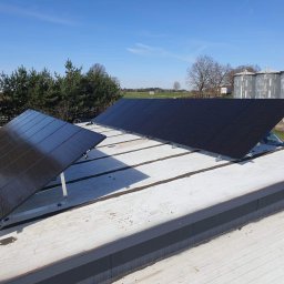 JLN Technology Systems Dariusz Zbigniew Urbański - Panele fotowoltaiczne zamontowane na płaskim dachu budynku przemysłowego, w tle silosy zbożowe i błękitne niebo.