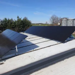 Panele fotowoltaiczne zamontowane na płaskim dachu budynku przemysłowego, w tle silosy zbożowe i błękitne niebo.