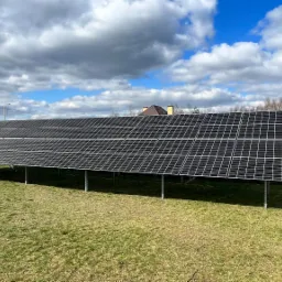 Instalacja fotowoltaiczna na metalowej konstrukcji, ustawiona na trawniku przed domem jednorodzinnym, widok w słoneczny dzień z chmurami.