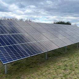 JLN Technology Systems Dariusz Zbigniew Urbański - Instalacja paneli fotowoltaicznych na trawniku w Turku, widoczne podpory i moduły pod kątem, pochmurne niebo w tle.