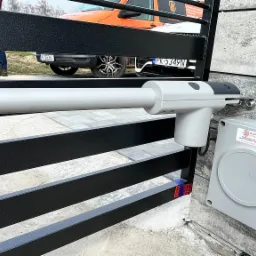 Szary siłownik bramy automatycznej Nice zamontowany na czarnej metalowej bramie posesji, z widocznym panelem sterującym i fragmentem pomarańczowego samochodu dostawczego w tle.