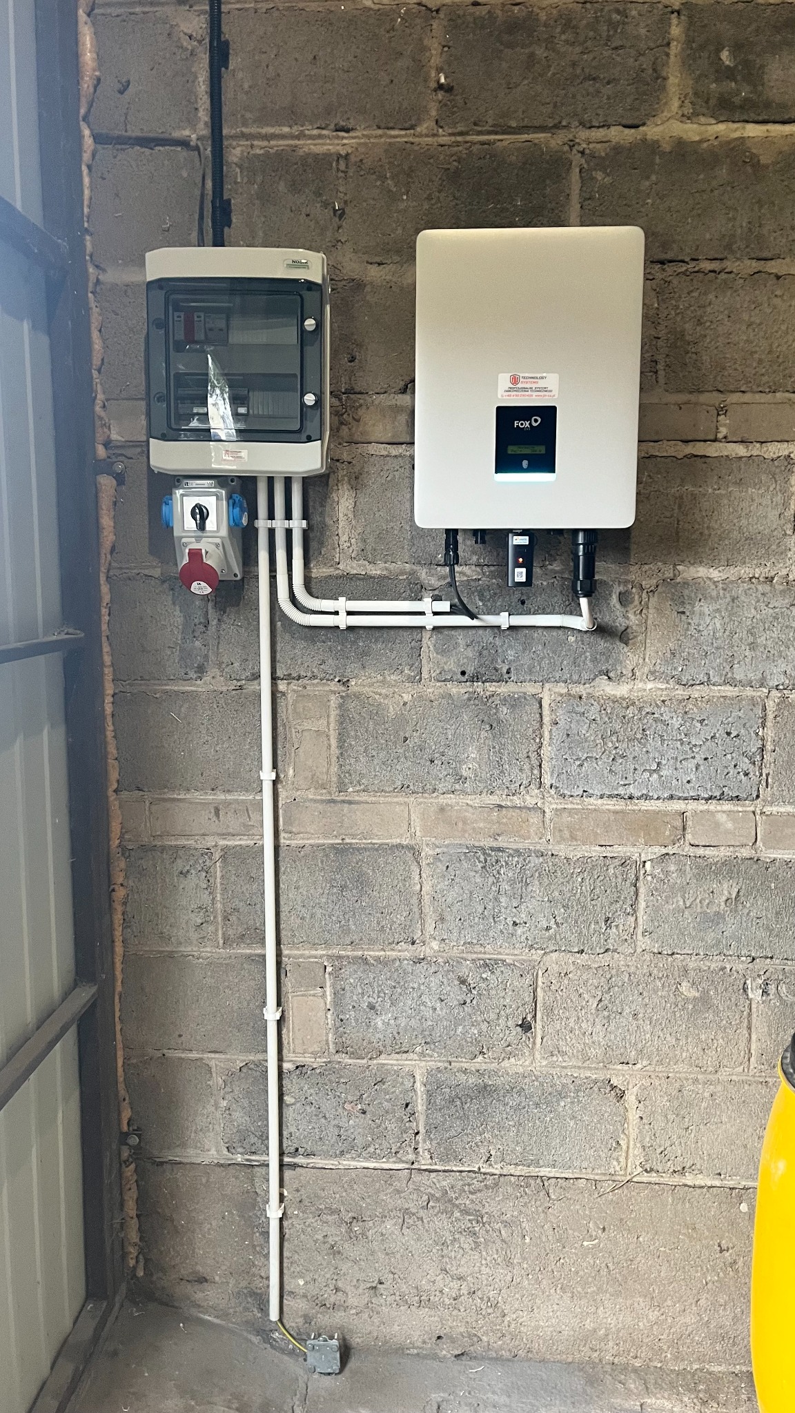 Rozdzielnia elektryczna i inwerter fotowoltaiczny zamontowane na szarej, betonowej ścianie z widocznymi przewodami w białych rurkach PCV.