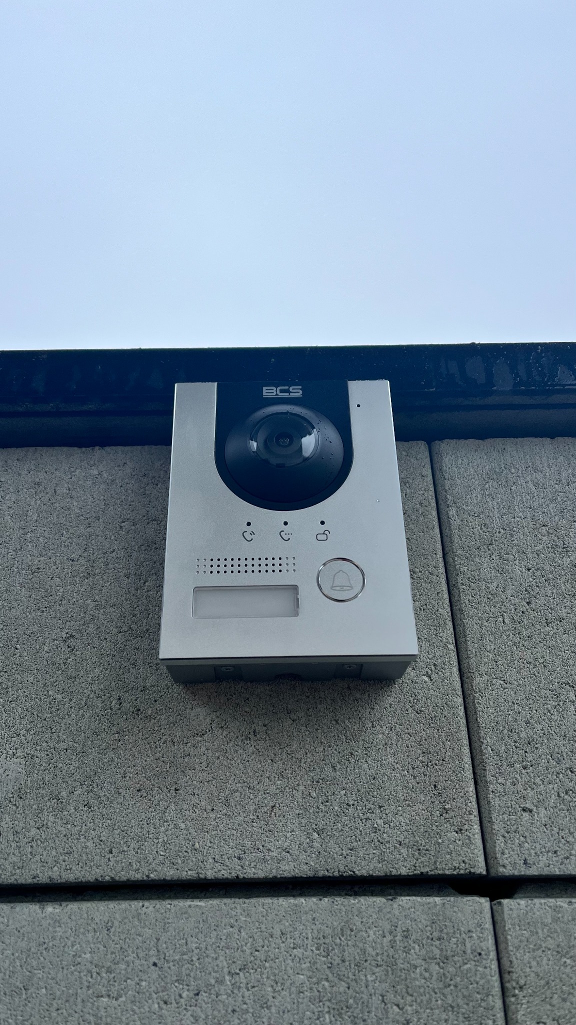 Srebrny wideodomofon BCS zamontowany na szarej, betonowej ścianie zewnętrznej budynku, widok z dołu.