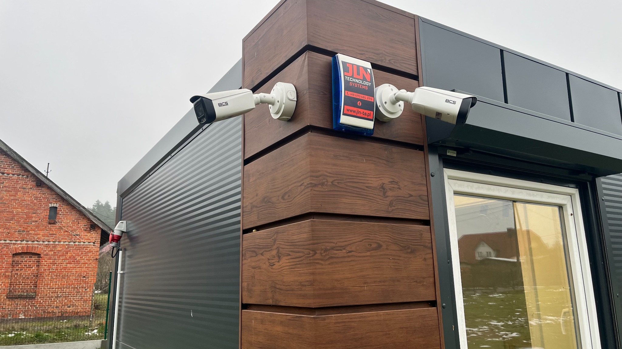 Dwie kamery monitoringu BCS zamontowane na rogu budynku z drewnopodobną elewacją, obok widoczna tabliczka informacyjna firmy JLN Technology Systems i fragment budynku z oknem.