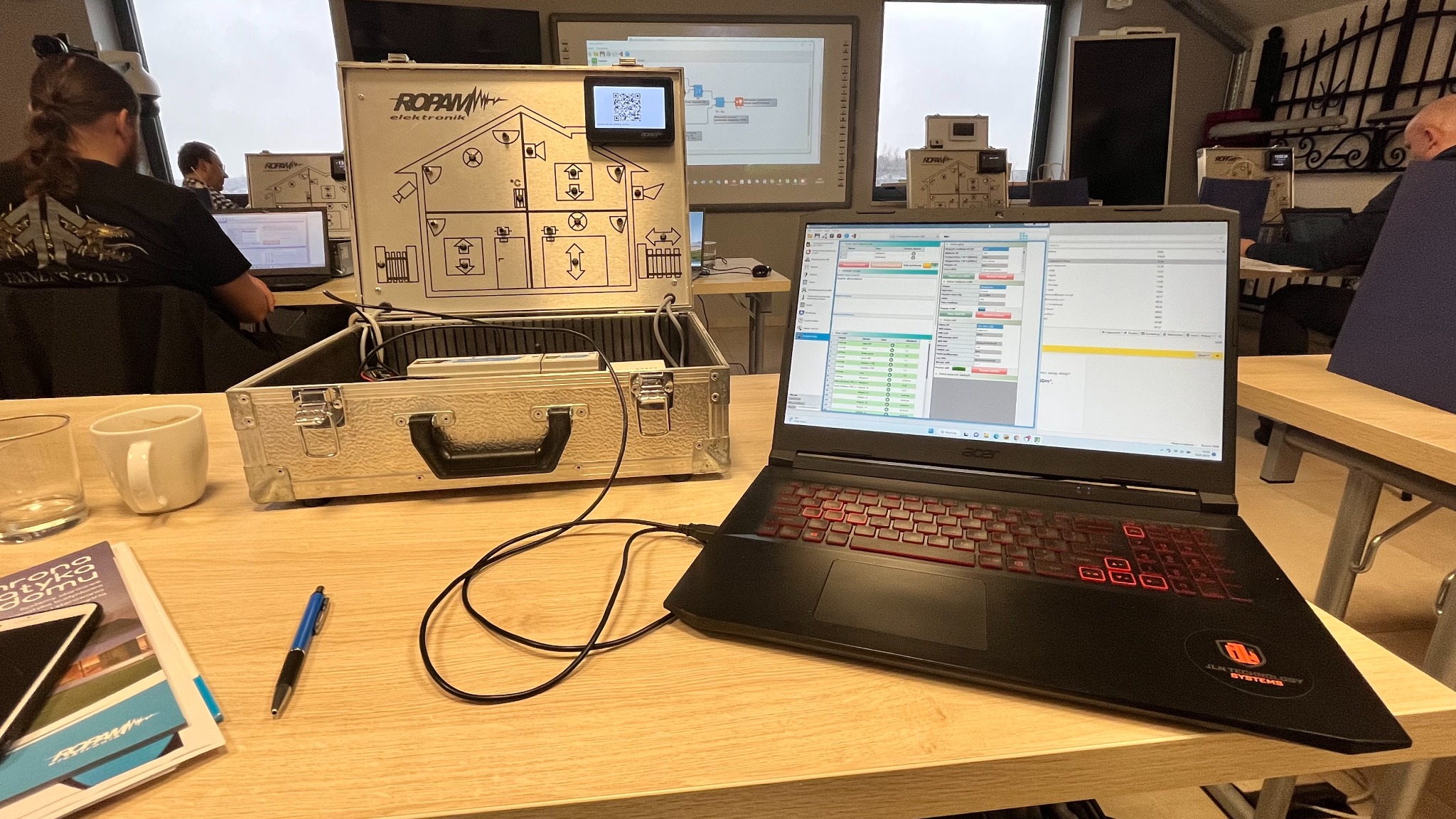 Laptop z uruchomionym oprogramowaniem do konfiguracji systemu alarmowego, obok walizka z panelem sterującym i schematem instalacji, widoczni ludzie w tle.