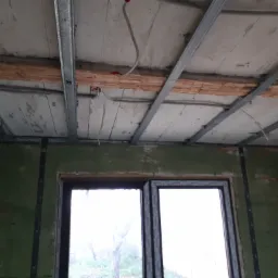 Wnętrze w trakcie prac: metalowy stelaż sufitowy przymocowany do drewnianych belek, zielone ściany z widocznymi oknami, instalacja elektryczna w suficie.