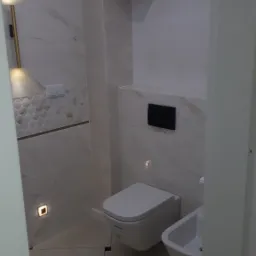 Nowoczesna łazienka z wiszącą toaletą i bidetem, wykończona białymi płytkami imitującymi marmur z delikatnym złotym żyłowaniem, oświetlona subtelnymi punktowymi lampami podłogowymi i listwą LED...