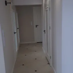 Korytarz w jasnych barwach z kremową podłogą w kwadraty z czarnymi wstawkami, białymi drzwiami z czarnymi klamkami i kinkietem w stylu retro z kloszem z przezroczystego szkła.