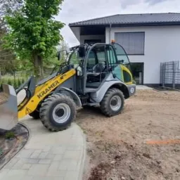 Żółto-szara ładowarka Kramer z pełną łyżką żwiru na tle nowoczesnego domu i dwóch pracowników w zielonych koszulkach, jeden z łopatą, obok ułożona kostka brukowa i rozłożona agrowłóknina.