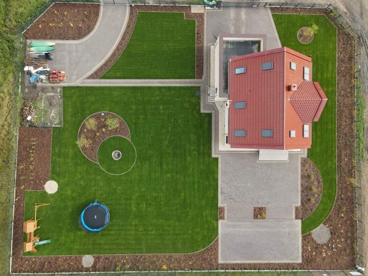 Widok z góry na zadbany ogród z równo przystrzyżonym trawnikiem, ścieżkami z kostki brukowej, placem zabaw z trampoliną i drewnianym domkiem, oraz obrzeżami z kory drzewnej.