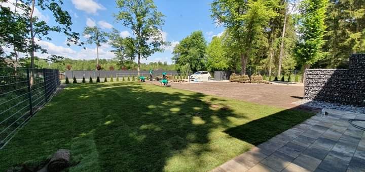 Układanie trawnika z rolki na posesji z ogrodzeniem z siatki i gabionami, widoczni pracownicy w zielonych strojach i zaparkowany samochód.