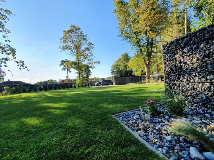 Starannie utrzymany trawnik z dekoracyjnym gabionem wypełnionym kamieniami w słoneczny dzień, widoczny fragment rabaty z kamykami i roślinami, w tle zaparkowany samochód.