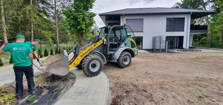 Żółto-szara ładowarka Kramer z pełną łyżką żwiru na tle nowoczesnego domu i dwóch pracowników w zielonych koszulkach, jeden z łopatą, obok ułożona kostka brukowa i rozłożona agrowłóknina.
