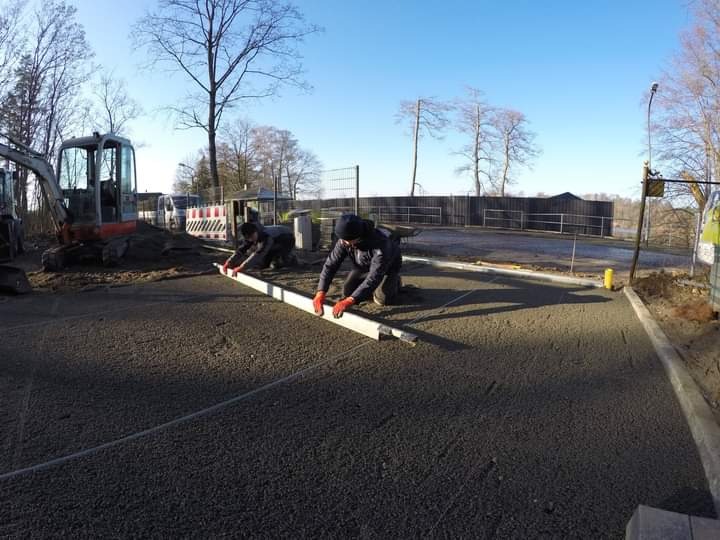 Dwóch pracowników w pomarańczowych rękawicach wyrównuje świeżo wylaną, ciemną nawierzchnię za pomocą długiej poziomicy. W tle widoczny minikoparka i ogrodzenie.