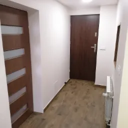 Korytarz po remoncie: drewnopodobne panele podłogowe, białe ściany, grzejnik, dwoje drzwi wewnętrznych - jedne z przeszkleniami, drugie pełne.