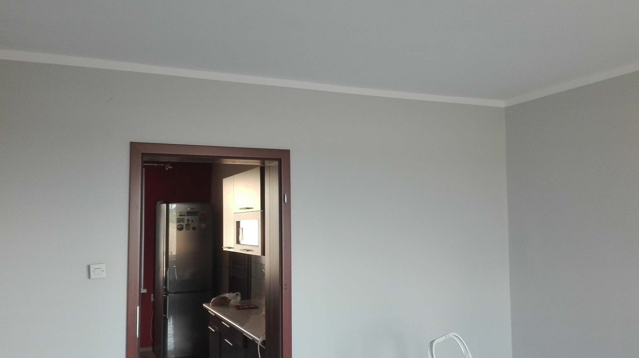 Świeżo pomalowane na szaro ściany z białą listwą przypodłogową i brązową futryną drzwiową, widok na kuchnię z lodówką i szafkami.