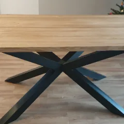 Stół z jasnym drewnianym blatem i czarną, geometryczną podstawą w kształcie gwiazdy, ustawiony na drewnianej podłodze, z choinką w tle.