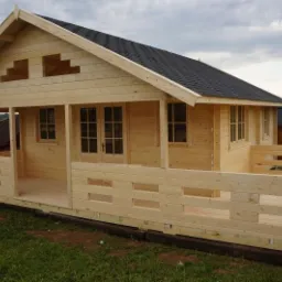 Surowy, drewniany domek letniskowy z czarnym dachem z gontu bitumicznego, częściowo zmontowany, z widocznymi elementami konstrukcyjnymi i pustymi przestrzeniami zamiast okien, na tle zielonej trawy...