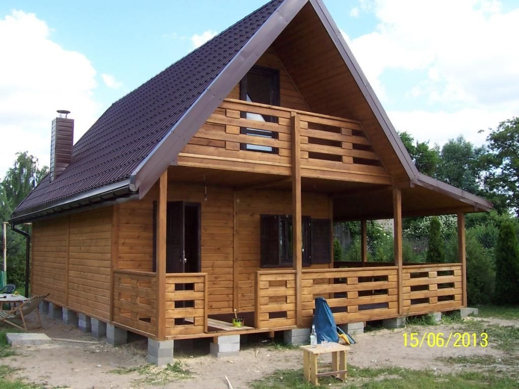 Gotowy, drewniany domek letniskowy z brązowym dachem i kominem, wzniesiony na betonowych bloczkach, z widoczną datą wykonania zdjęcia w prawym dolnym rogu.