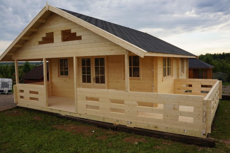 Surowy, drewniany domek letniskowy z czarnym dachem z gontu bitumicznego, częściowo zmontowany, z widocznymi elementami konstrukcyjnymi i pustymi przestrzeniami zamiast okien, na tle zielonej trawy...