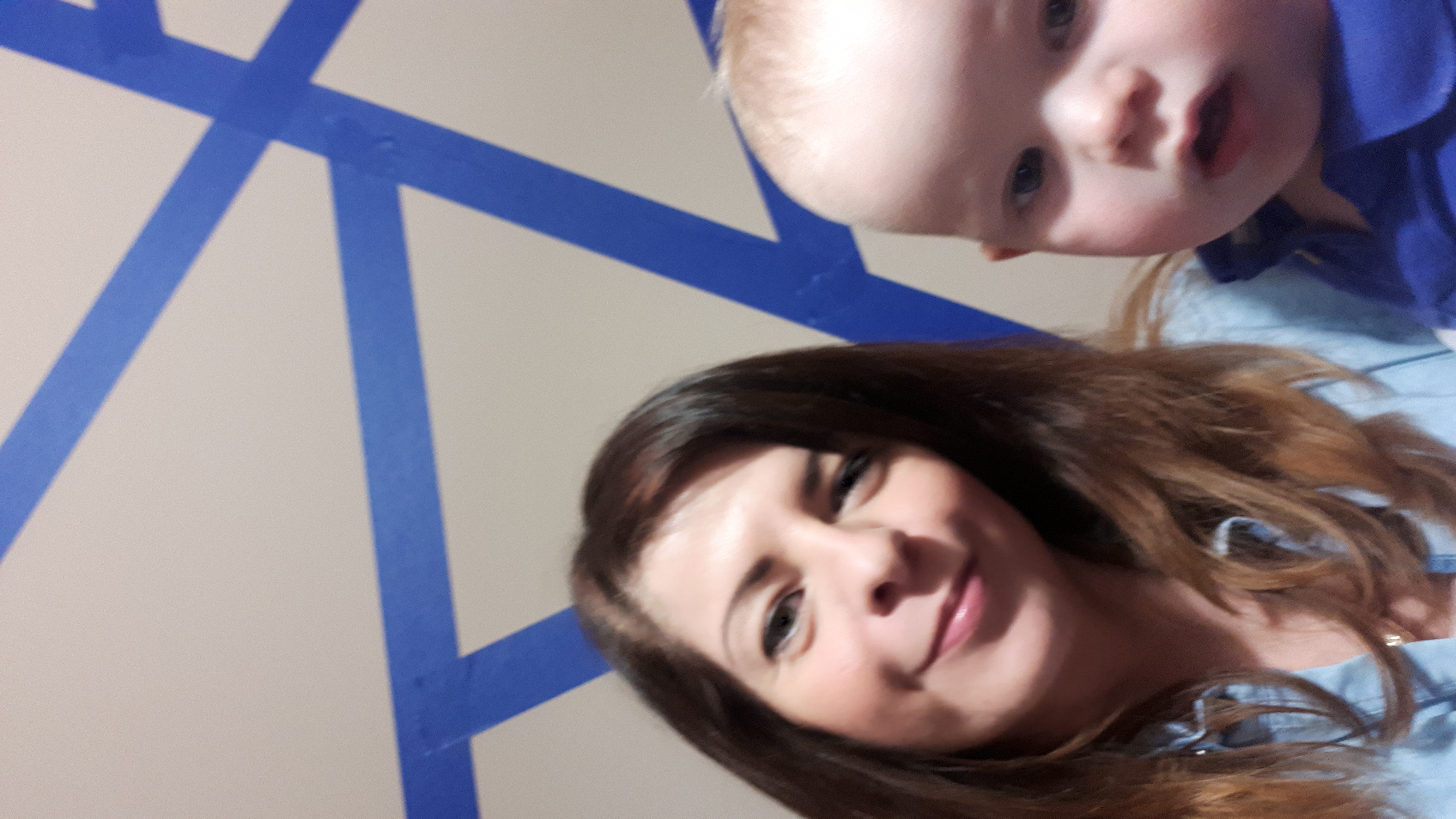 Kobieta z ciemnymi włosami i małe dziecko w niebieskich ubraniach pozują do zdjęcia na tle ściany z niebieskimi paskami taśmy malarskiej.